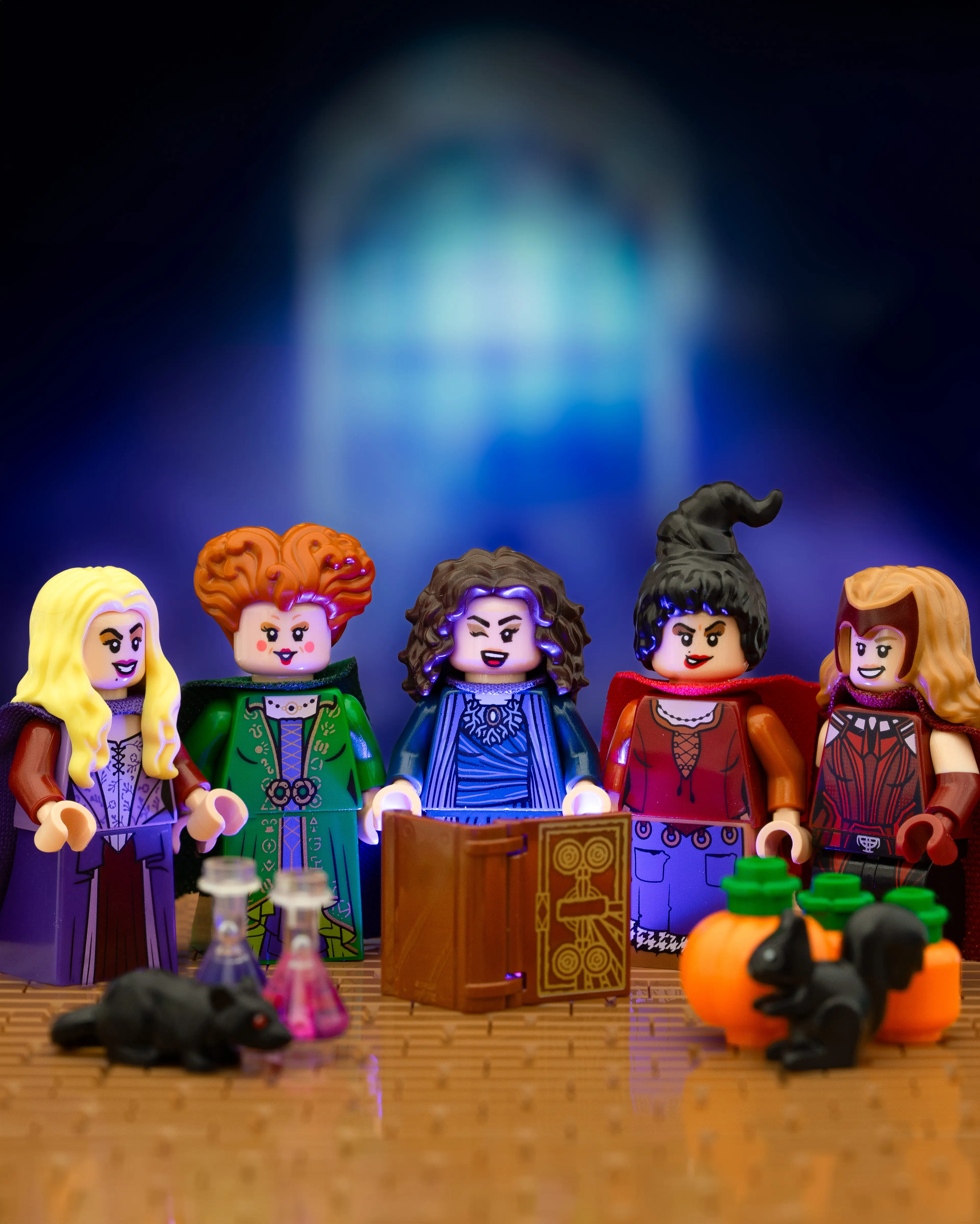 Five LEGO witches gather around an ancient spellbook glowing with eerie purple light, surrounded by potions, pumpkins, and black animals, set against a mystical arched window backdrop.